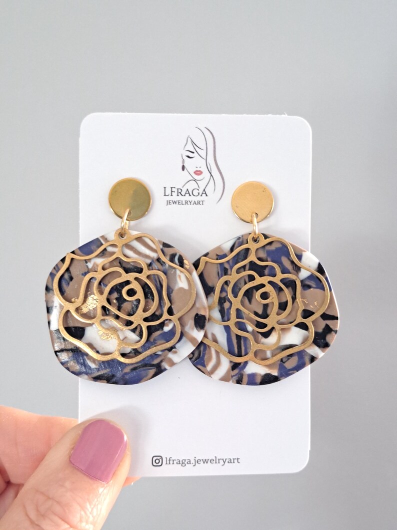 May include: A pair of statement earrings featuring a floral design. The earrings have a gold-colored metal overlay on a marbled base of white, navy, and brown. The earrings are displayed on a white card with the text "LFRAGA JEWELRYART".