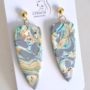 May include: A pair of handmade earrings with a marbled design in shades of gray, teal, and gold. The earrings have a teardrop shape and are suspended from gold-colored ball studs. The earrings are displayed on a white card with the text "LFRAGA JEWELRYART".