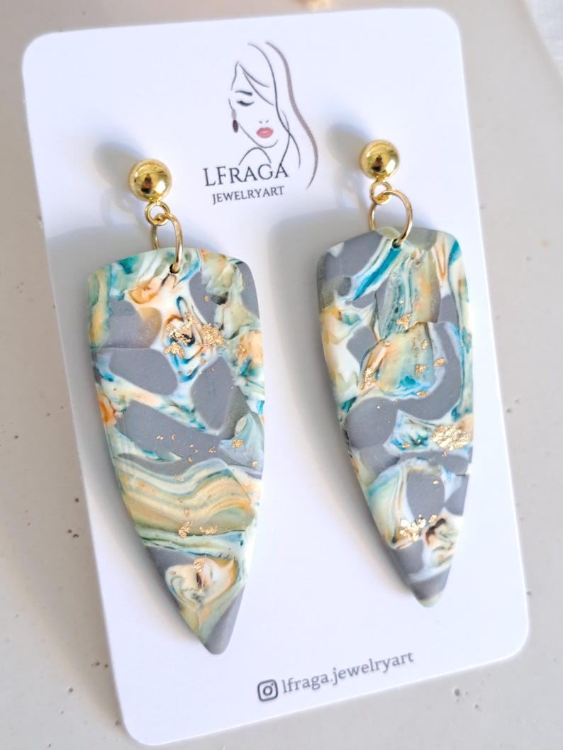 May include: A pair of handmade earrings with a marbled design in shades of gray, teal, and gold. The earrings have a teardrop shape and are suspended from gold-colored ball studs. The earrings are displayed on a white card with the text "LFRAGA JEWELRYART".
