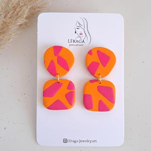 Pode incluir: Um par de brincos abstratos laranja e rosa. Os brincos são feitos de um material liso e apresentam um gancho dourado. Os brincos são exibidos em um cartão branco com o nome e o logotipo da marca.