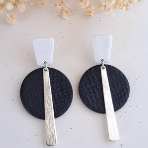 Peut inclure: Paire de boucles d'oreilles modernes. Elles sont composées d'un carré blanc texturé, d'un cercle noir texturé et d'une barre argentée. Les boucles d'oreilles sont présentées sur un fond gris clair, avec des fleurs séchées.