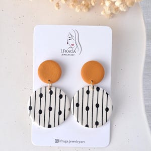 Puede incluir: Un par de pendientes colgantes con un patrón geométrico blanco y negro y una pequeña parte superior redonda de color amarillo mostaza. Los pendientes se muestran en una tarjeta blanca con el texto "LFRAGA JEWELRYART" y el nombre de usuario de Instagram "@lfraga.jewelryart".