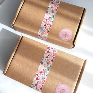 May include: Two brown cardboard boxes with floral patterned tape wrapped around them. Each box has a pink circular sticker with the words "THANK YOU" and a floral design. The boxes are on a white surface.