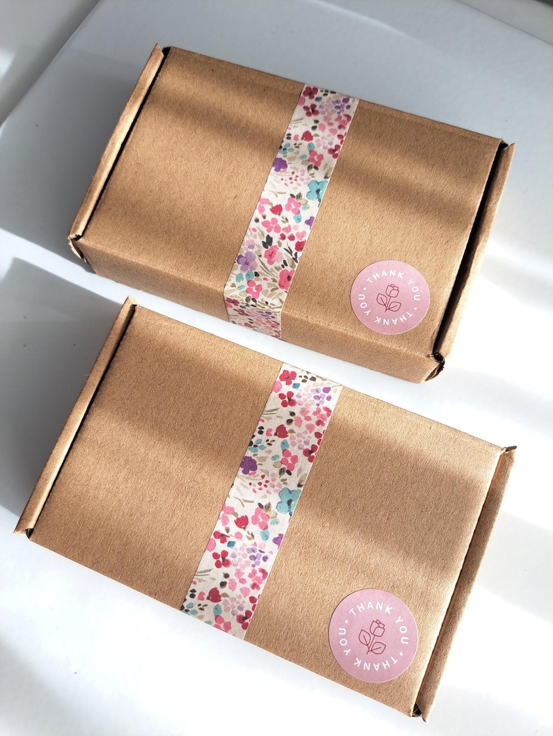 May include: Two brown cardboard boxes with floral patterned tape wrapped around them. Each box has a pink circular sticker with the words "THANK YOU" and a floral design. The boxes are on a white surface.