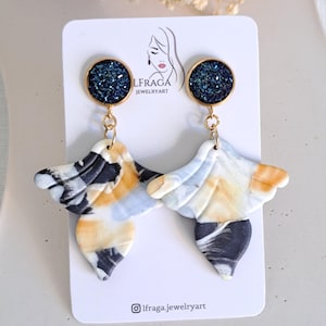 May include: A pair of handmade earrings with a unique design. Each earring features a dark blue, glittery circular top and a marbled, wing-shaped bottom in shades of black, white, and yellow. The earrings are displayed on a white card with the text "LFRAGA JEWELRYART".