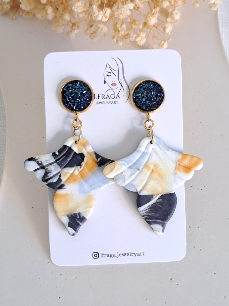 May include: A pair of handmade earrings with a unique design. Each earring features a dark blue, glittery circular top and a marbled, wing-shaped bottom in shades of black, white, and yellow. The earrings are displayed on a white card with the text "LFRAGA JEWELRYART".