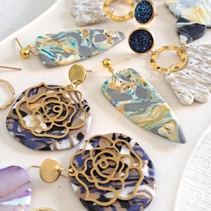 May include: A collection of handmade earrings in various designs. Some earrings feature marbled polymer clay in shades of gray, blue, yellow, and white. Others have gold-colored metal rose cutouts or circular accents with dark blue glitter.