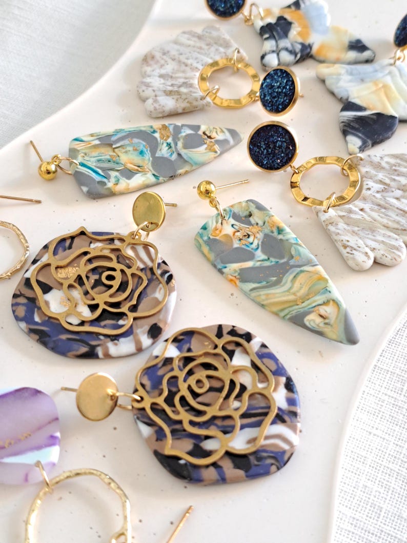 May include: A collection of handmade earrings in various designs. Some earrings feature marbled polymer clay in shades of gray, blue, yellow, and white. Others have gold-colored metal rose cutouts or circular accents with dark blue glitter.