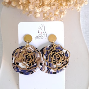May include: A pair of statement earrings featuring a gold-colored rose design overlaying a marbled base in shades of white, brown, and blue. The earrings have a circular gold stud at the top and are displayed on a white card with the brand logo.