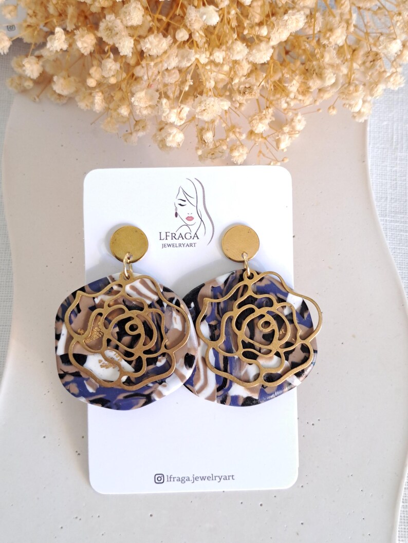 May include: A pair of statement earrings featuring a gold-colored rose design overlaying a marbled base in shades of white, brown, and blue. The earrings have a circular gold stud at the top and are displayed on a white card with the brand logo.