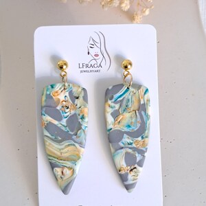 May include: A pair of handmade, marbled polymer clay earrings. The earrings are gray with swirls of blue, yellow, and gold. Each earring has a gold-colored stud and a teardrop shape. The earrings are displayed on a white card with the brand logo.