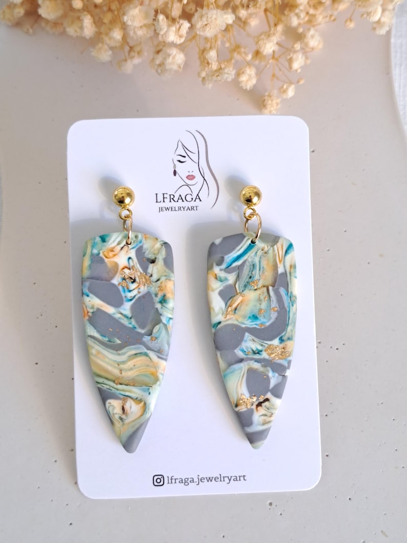 May include: A pair of handmade, marbled polymer clay earrings. The earrings are gray with swirls of blue, yellow, and gold. Each earring has a gold-colored stud and a teardrop shape. The earrings are displayed on a white card with the brand logo.
