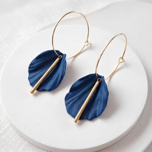 Op de afbeelding: Een paar oorringen met dunne, goudkleurige metalen hoepels. Elke oorbel is versierd met een donkerblauw, bloemblad-vormig onderdeel en een verticale goudkleurige metalen staaf die in het midden van het blauwe bloemblad-element rust.