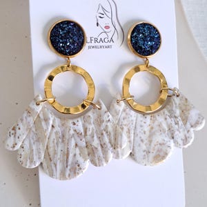 May include: A pair of dangle earrings with a gold-tone metal design. The earrings feature a dark blue, glittery circular top, a gold ring, and a white and gold marbled fan-shaped bottom. The earrings are displayed on a white card with the brand name and logo.