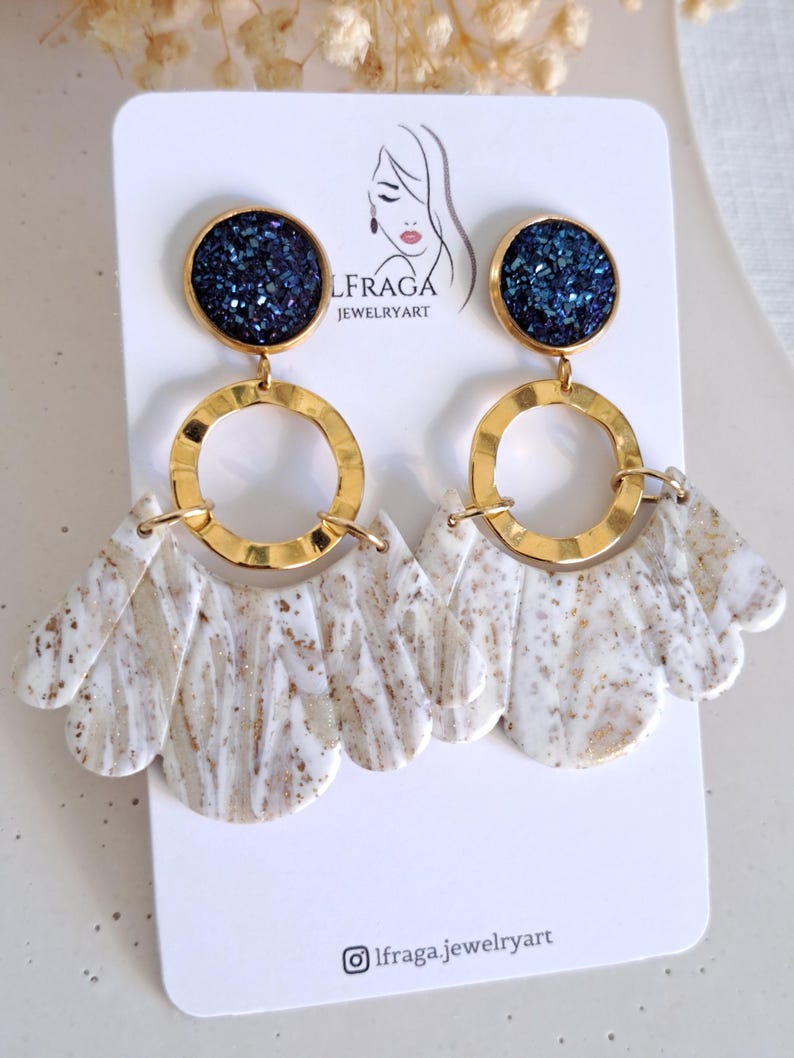 May include: A pair of dangle earrings with a gold-tone metal design. The earrings feature a dark blue, glittery circular top, a gold ring, and a white and gold marbled fan-shaped bottom. The earrings are displayed on a white card with the brand name and logo.