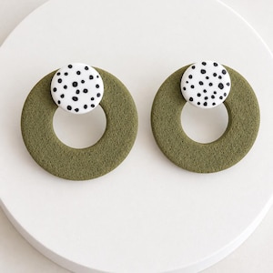 Pode incluir: Um par de brincos com um grande anel texturizado verde-oliva preso a um pequeno pino circular branco com um padrão de manchas pretas. Os brincos têm um acabamento fosco e um design geométrico, destacando o contraste entre a cor verde terrosa sólida e o detalhe de pontos brancos e pretos na parte superior.
