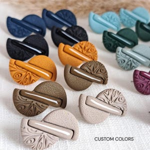 Può includere: Una collezione di orecchini a lobo semicircolari in vari colori, tra cui blu navy, nero, senape, marrone, beige, verde ottanio e viola. Ogni orecchino presenta un motivo testurizzato e una barra liscia e lucida. Il testo recita CUSTOM COLORS.
