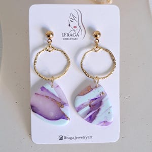 May include: A pair of handmade earrings featuring gold-colored hoops and abstract, marbled pendants in shades of purple, white, and light blue. The earrings are displayed on a white card with the brand name and logo.