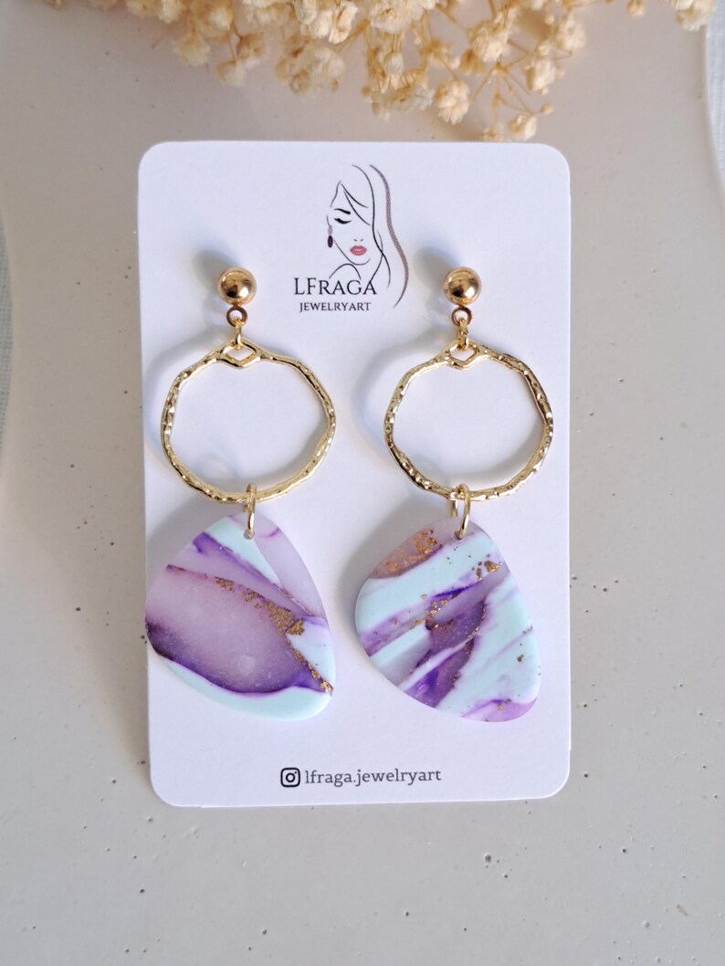 May include: A pair of handmade earrings featuring gold-colored hoops and abstract, marbled pendants in shades of purple, white, and light blue. The earrings are displayed on a white card with the brand name and logo.