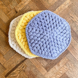 May include: Three handmade crochet coasters in a row. The coasters are in shades of cream, yellow, and light blue. The coasters are round with a textured, raised pattern. The coasters are on a wooden surface.