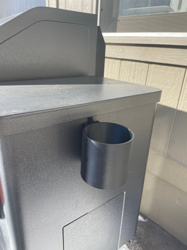 Puede incluir: Un portavasos de pl&aacute;stico negro fijado a un buz&oacute;n gris.