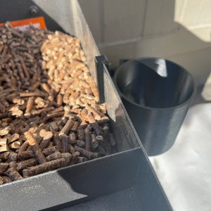 Puede incluir: Una parrilla de pellets de metal negro con un portavasos de pl&aacute;stico negro. La parrilla est&aacute; llena de pellets de madera.