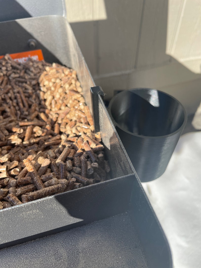 Puede incluir: Una parrilla de pellets de metal negro con un portavasos de pl&aacute;stico negro. La parrilla est&aacute; llena de pellets de madera.