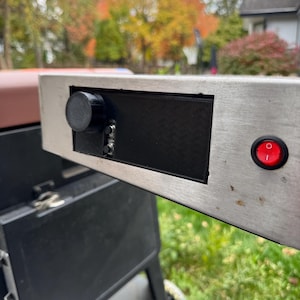 May include: A stainless steel control panel with a black knob, a black rectangular section, and a red illuminated power switch. The panel is attached to a black grill, with a blurred outdoor background.