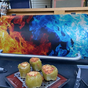 May include: A black grill with a flame design on the lid, with yellow, orange, and blue colours. Four green stuffed bell peppers sit on a metal rack. Tongs and a knife are placed nearby.