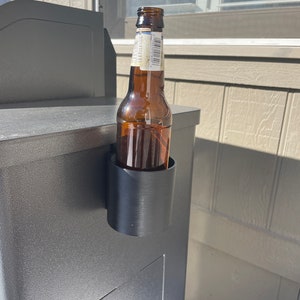 Puede incluir: Un porta botellas de pl&aacute;stico negro fijado a una superficie gris, sosteniendo una botella de vidrio marr&oacute;n.