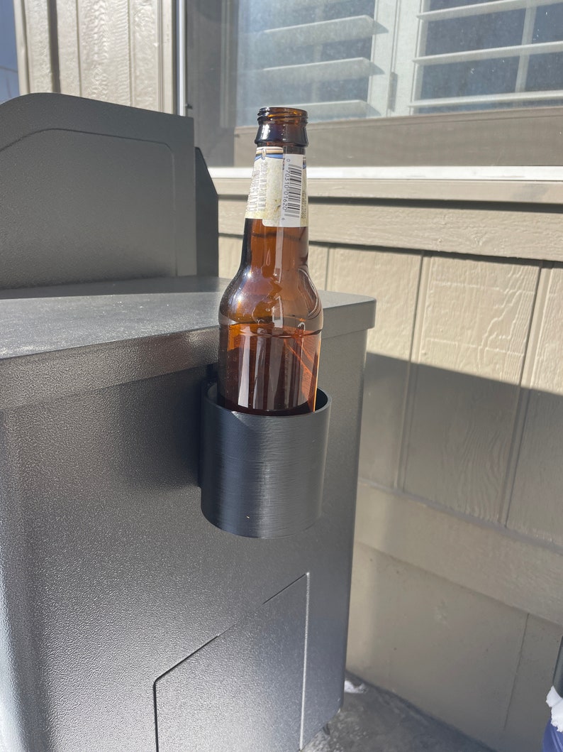 Puede incluir: Un porta botellas de pl&aacute;stico negro fijado a una superficie gris, sosteniendo una botella de vidrio marr&oacute;n.