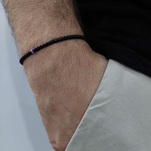 May include: Black beaded bracelet with a small purple accent. The bracelet is worn on a wrist, with a black shirt and light-colored pants in the background. The bracelet appears to be made of small, square beads.