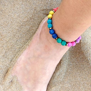May include: A colorful beaded anklet with yellow, orange, red, pink, blue, and green beads. The anklet is on a person's ankle, with the foot resting on sand.