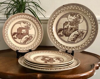 May include: A collection of vintage brown and cream-colored plates with detailed landscape and fan designs. The plates are displayed on a dark wooden table, showcasing a variety of sizes and patterns. A decorative plant is in the background.