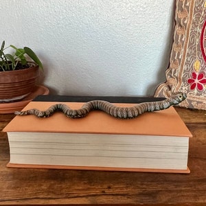 May include: A brown book with a black spine and a gray and brown snake bookmark on top of it.