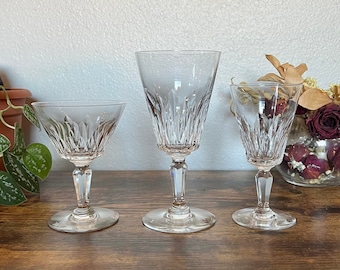 Op de afbeelding: Drie heldere kristallen wijnglazen met ingewikkelde verticale snijpatronen. De glazen hebben verschillende maten, met een steelontwerp en ronde bodems. Ze staan op een donker houten oppervlak, met een plant en een decoratieve vaas op de achtergrond.