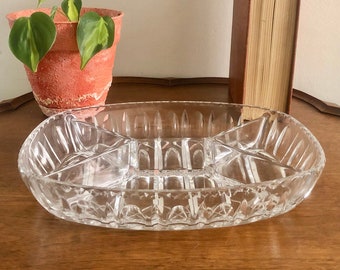 May include: Clear glass serving dish with multiple compartments. The oval-shaped dish has a textured, geometric pattern and is designed for serving snacks or appetizers. A small potted plant and a book are in the background.