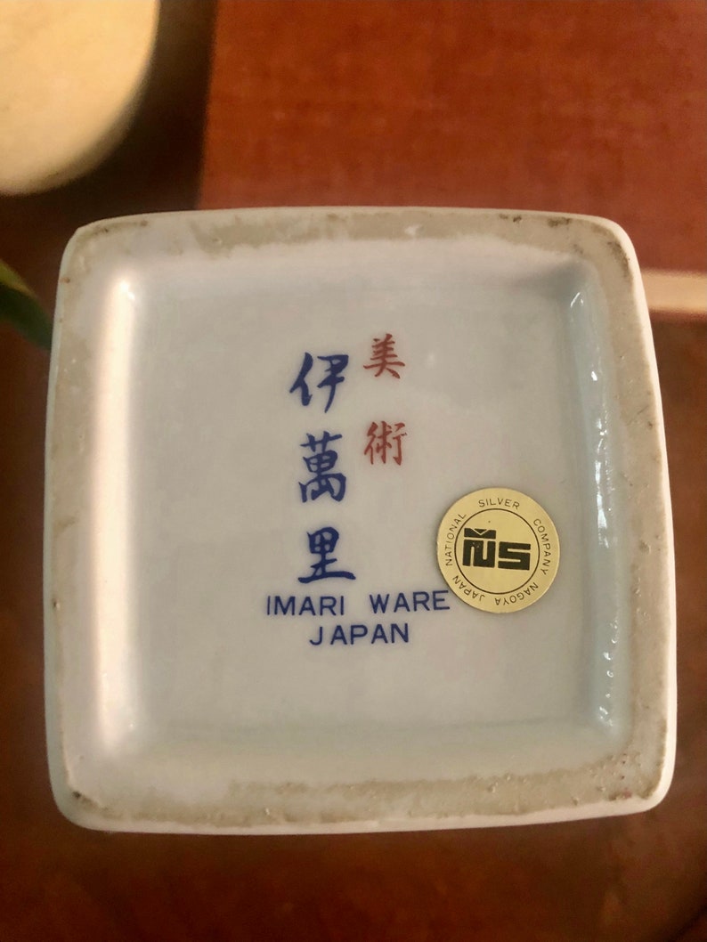 May include: A white Imari Ware ceramic piece with blue and red Japanese characters and the text "IMARI WARE JAPAN". A gold and white sticker reads "NATIONAL SILVER COMPANY NAGOYA".
