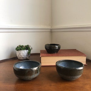May include: Three small ceramic bowls, two with a blue glaze and one with a black glaze. The bowls are sitting on a wooden table with a book and a small plant in a white pot.