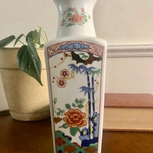 May include: A white ceramic vase with a rectangular body and a flared neck. The vase is decorated with floral and bamboo motifs in shades of red, blue, green, and gold. The vase is approximately 8 inches tall.