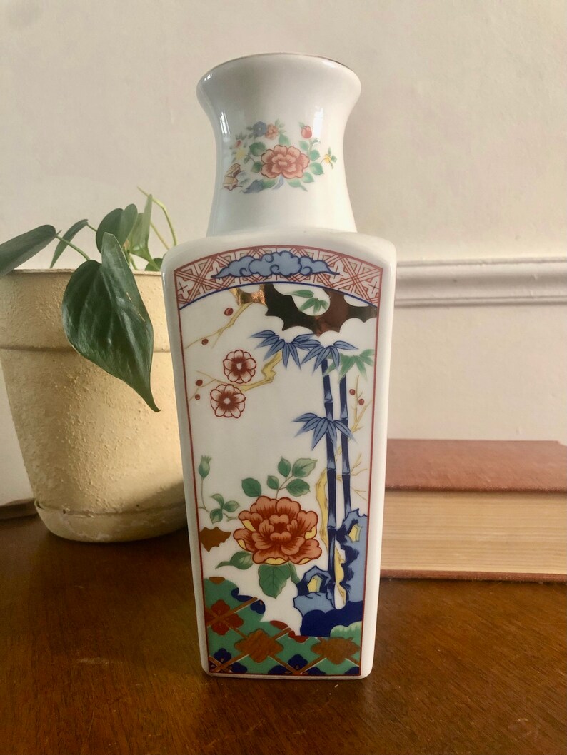 May include: A white ceramic vase with a rectangular body and a flared neck. The vase is decorated with floral and bamboo motifs in shades of red, blue, green, and gold. The vase is approximately 8 inches tall.