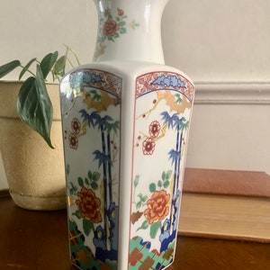 May include: A white porcelain vase with a hexagonal body and a flared neck. The vase is decorated with a floral and bamboo design in shades of red, blue, green, and gold. The rim is trimmed with gold.