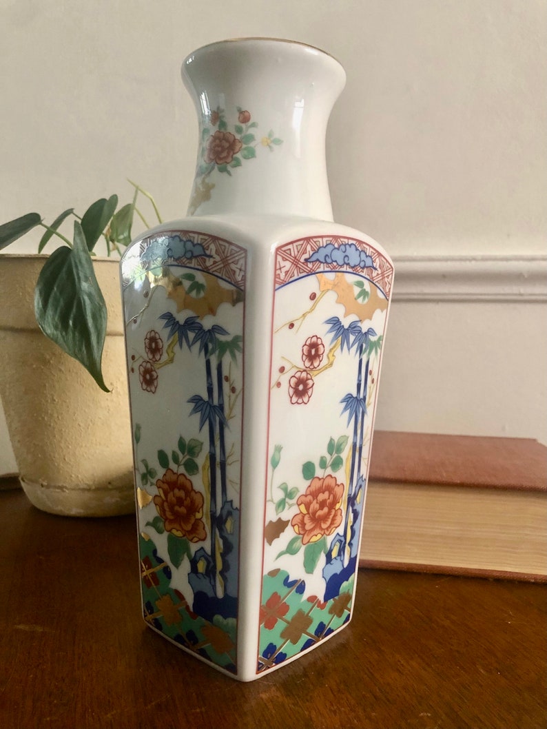 May include: A white porcelain vase with a hexagonal body and a flared neck. The vase is decorated with a floral and bamboo design in shades of red, blue, green, and gold. The rim is trimmed with gold.