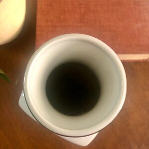May include: A white ceramic vase with a flared rim and a square base. The interior is dark, and the rim is accented with a thin brown line. The vase sits on a wooden surface, with a book and a decorative object in the background.