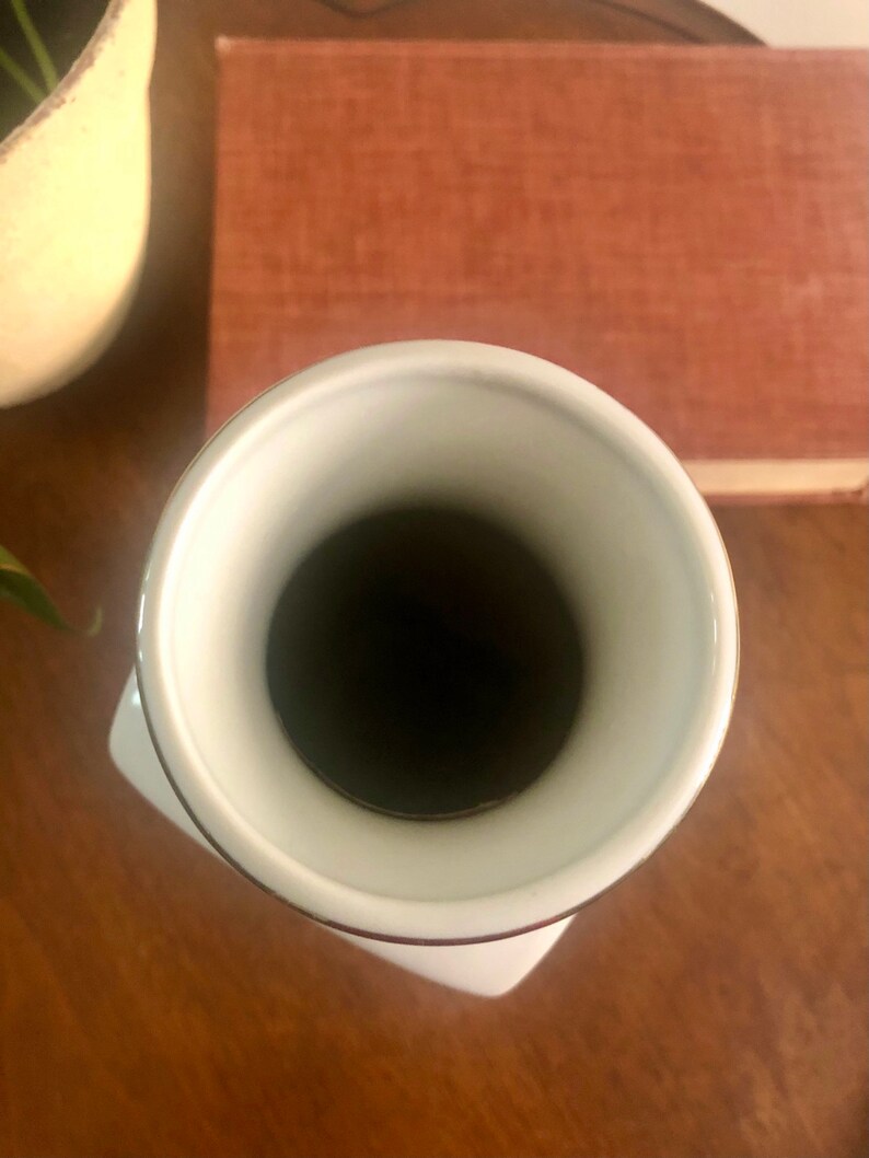 May include: A white ceramic vase with a flared rim and a square base. The interior is dark, and the rim is accented with a thin brown line. The vase sits on a wooden surface, with a book and a decorative object in the background.