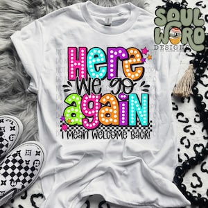 May include: White t-shirt with a colorful graphic that says "Here we go again I mean welcome back!" The graphic is in a retro style with a checkered background and stars.