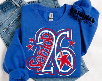 Puede incluir: Una sudadera azul real con la palabra "Senior" en rojo y blanco, y el número "26". El diseño incluye estrellas rojas y contornos blancos. La sudadera está colocada sobre un par de vaqueros azules.