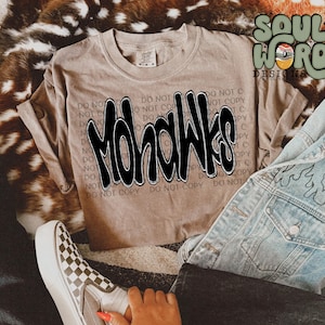 Puede incluir: Una camiseta marrón con la palabra "Mohawks" impresa en negro en la parte delantera. La camiseta está sobre una superficie de piel marrón con un par de zapatos Vans a cuadros y una chaqueta vaquera azul.