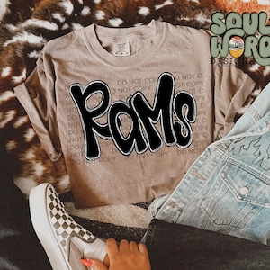 May include: A brown t-shirt with the word "Rams" written in black, outlined in white, in a graffiti style font. The shirt is on a brown fur surface with a pair of blue jeans and a pair of checkered Vans shoes.