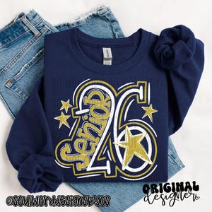 May include: A navy blue sweatshirt featuring the text "Senior 26" in gold and white, with gold star accents. The sweatshirt is placed on top of blue denim jeans. The words "Original Designer" are also visible.